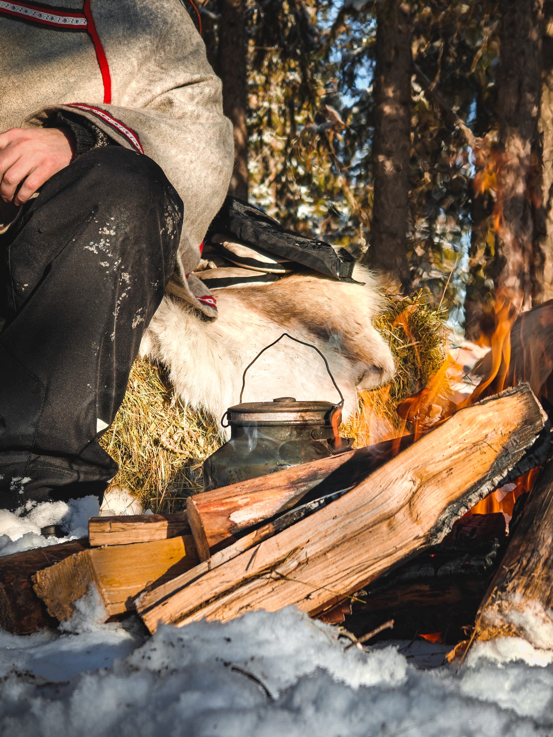 Contact and FAQ's page picture. A traditional Lapland campfire with water boiling in a soot-blackened kettle. In the background, a reindeer herder sits on top of a hay bale and reindeer skin, enjoying the sunny weather.