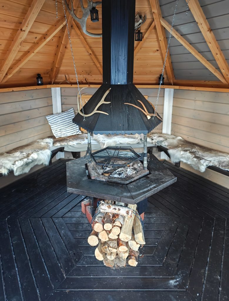 A picture of a traditional kota, with a campfire in the center where food can be prepared and enjoyed. In the foreground, firewood is stacked, and along the edges of the kota, you can sit on reindeer hides and relax.