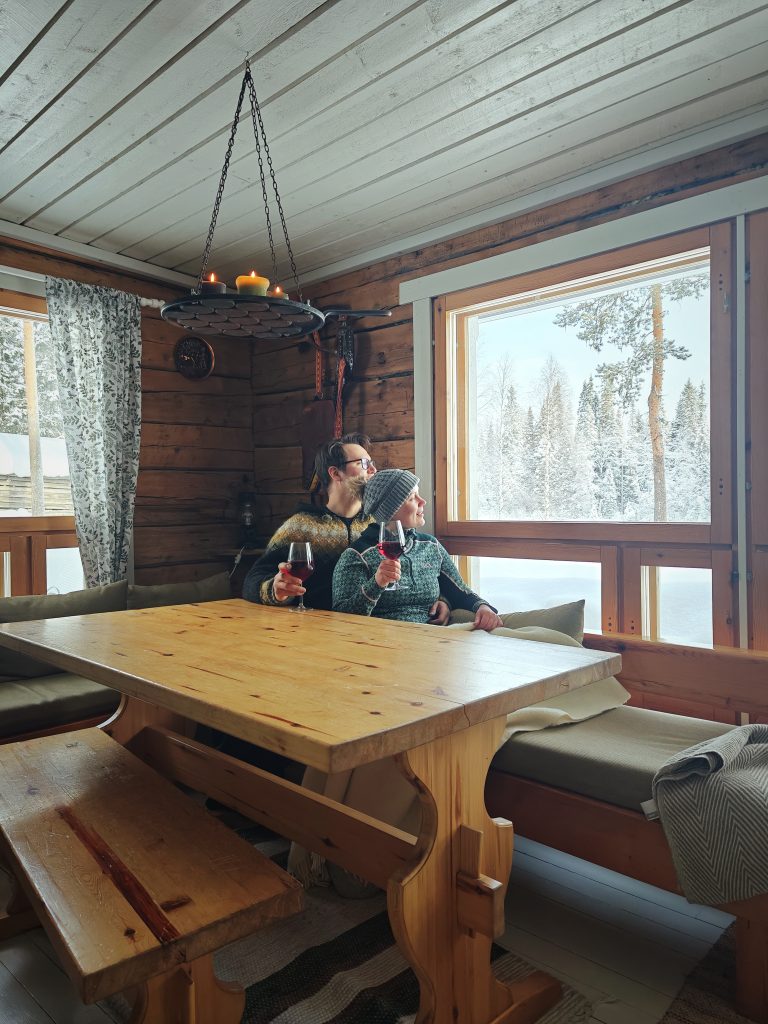 A young couple enjoying a drink, sitting at the dining table in the Kentura guesthouse. They are looking out at the stunning snowy riverside landscape visible through the window.