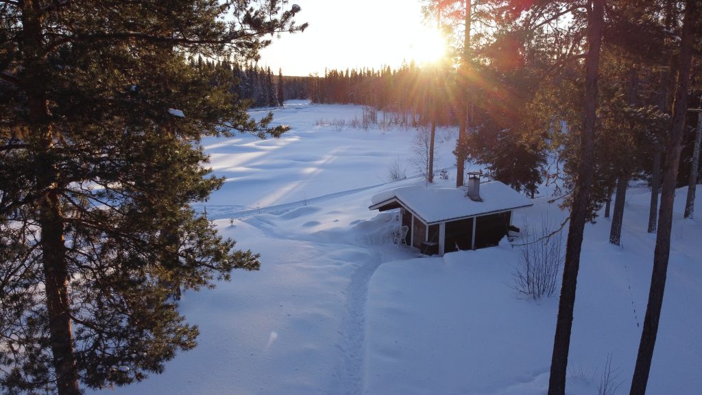 The Kentura Reindeer Farm A lakeside sauna by the river, with a snowy landscape and the sun low on the horizon. The river is frozen, and a small path leads to the sauna.
