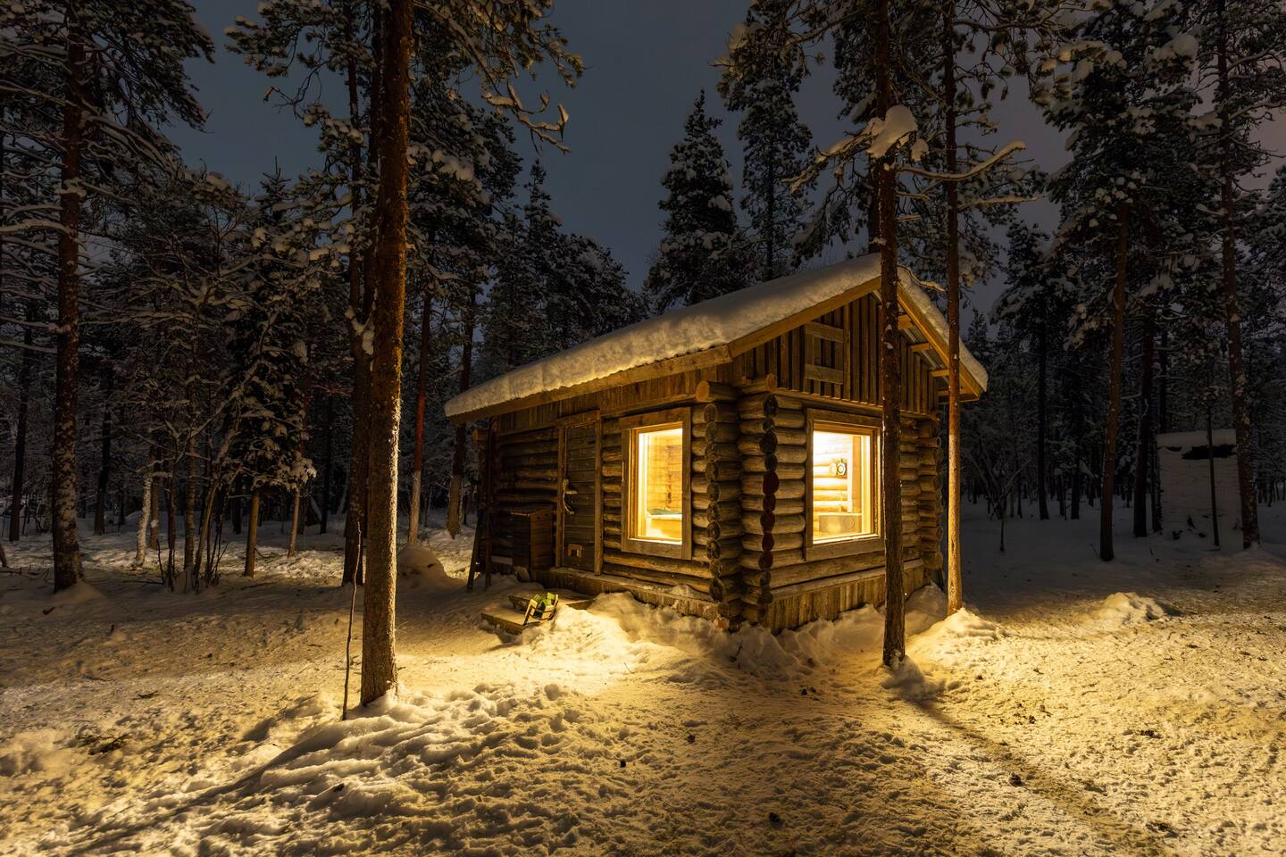 A dark, snowy setting inside a reindeer enclosure. The warm light from the Kentura cabin illuminates the snowy yard outside.